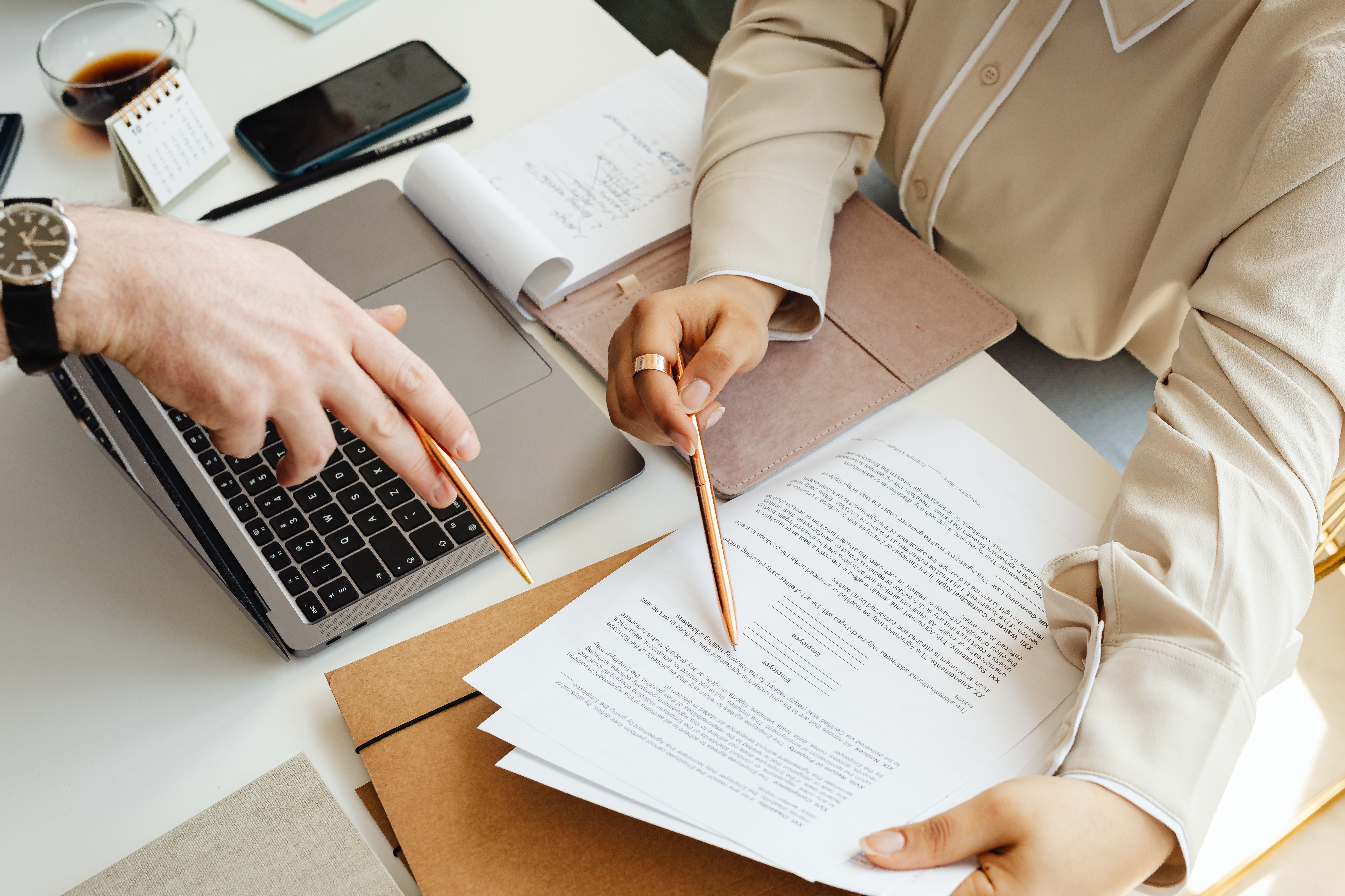 Duas pessoas comparando propostas em documentos com caneta e notebook sobre a mesa