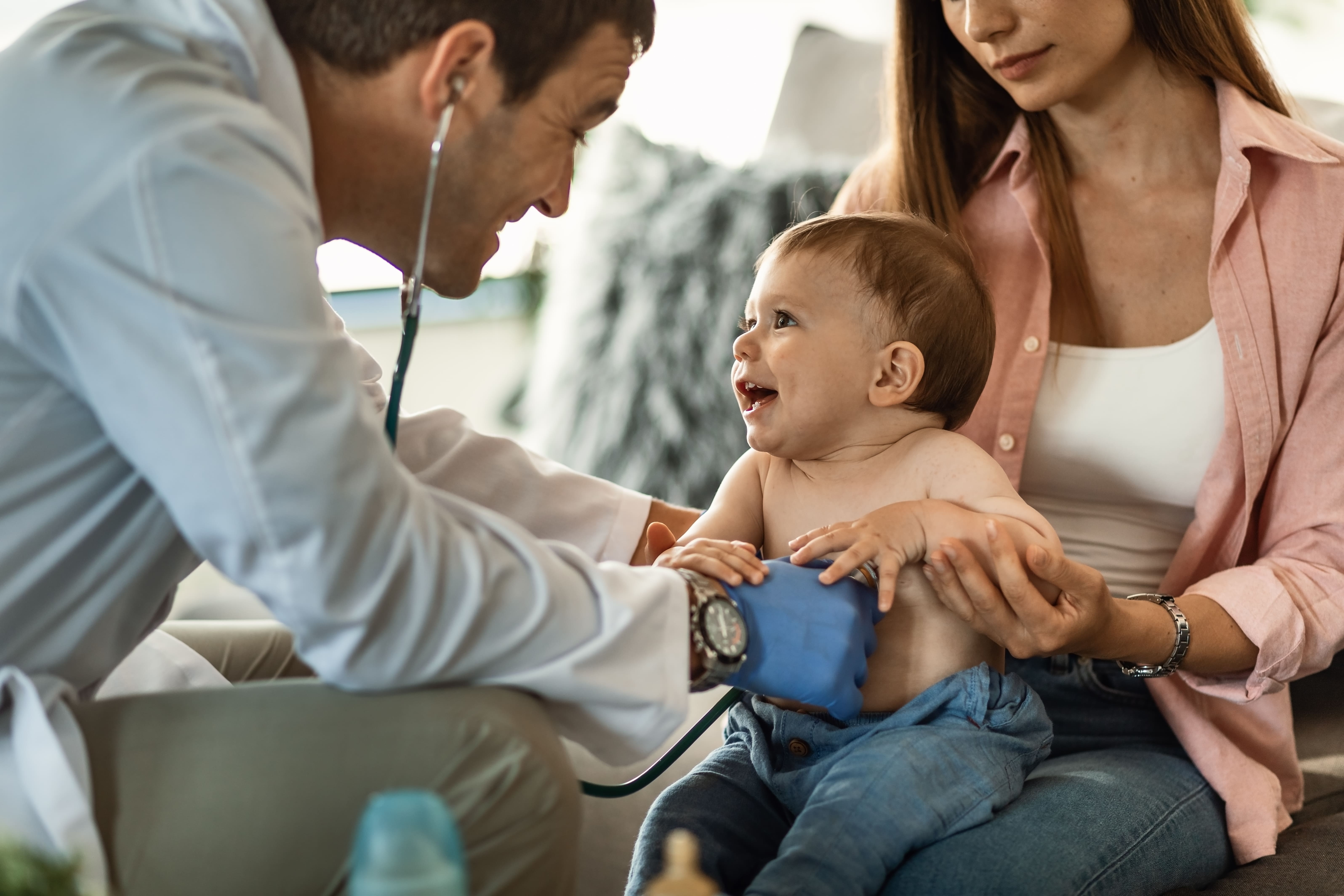 Pediatra sorridente examina um bebê sentado no colo da mãe durante uma consulta pediátrica.