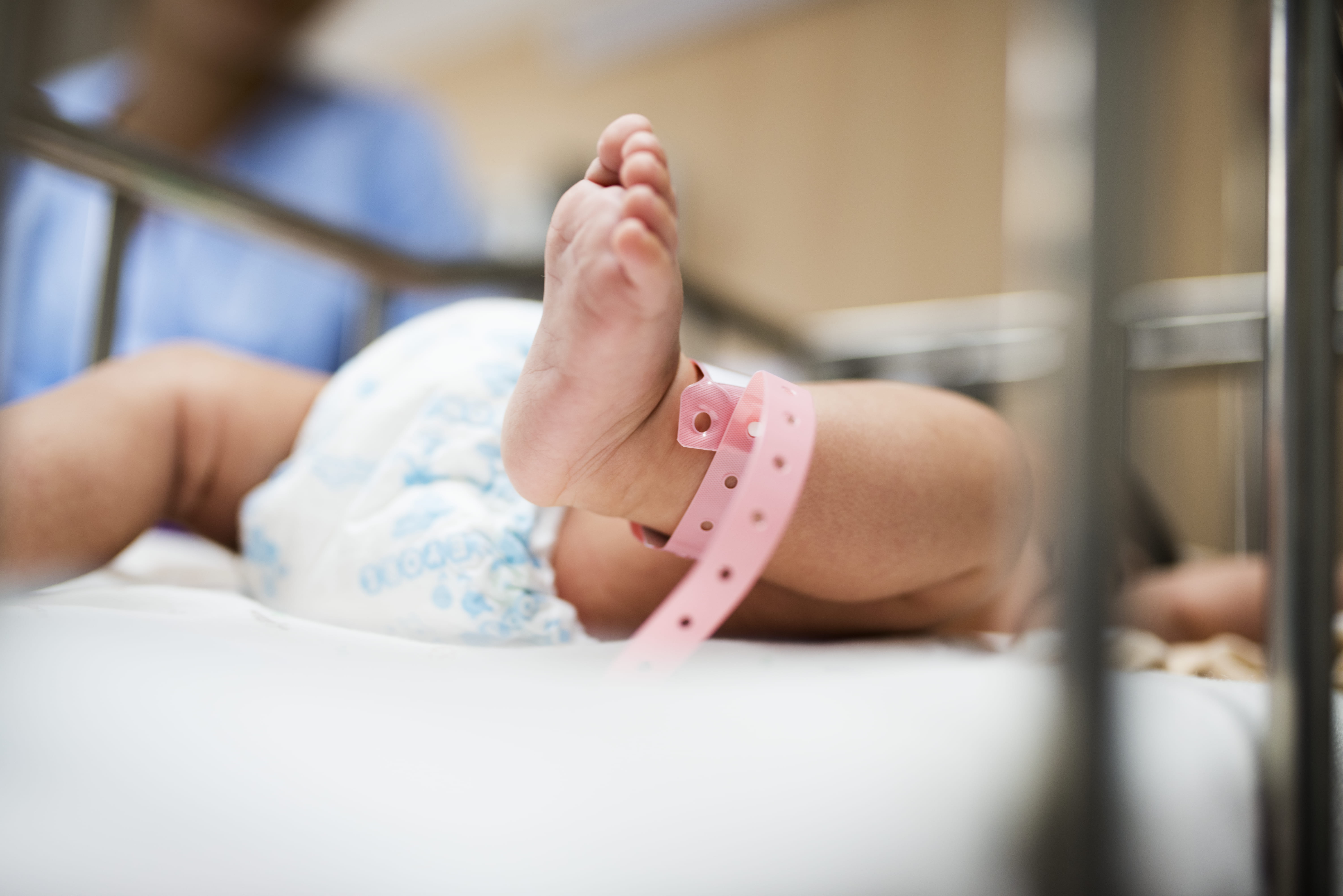 Pé de recém-nascido com pulseira de identificação em berço de maternidade, com profissional de saúde ao fundo.