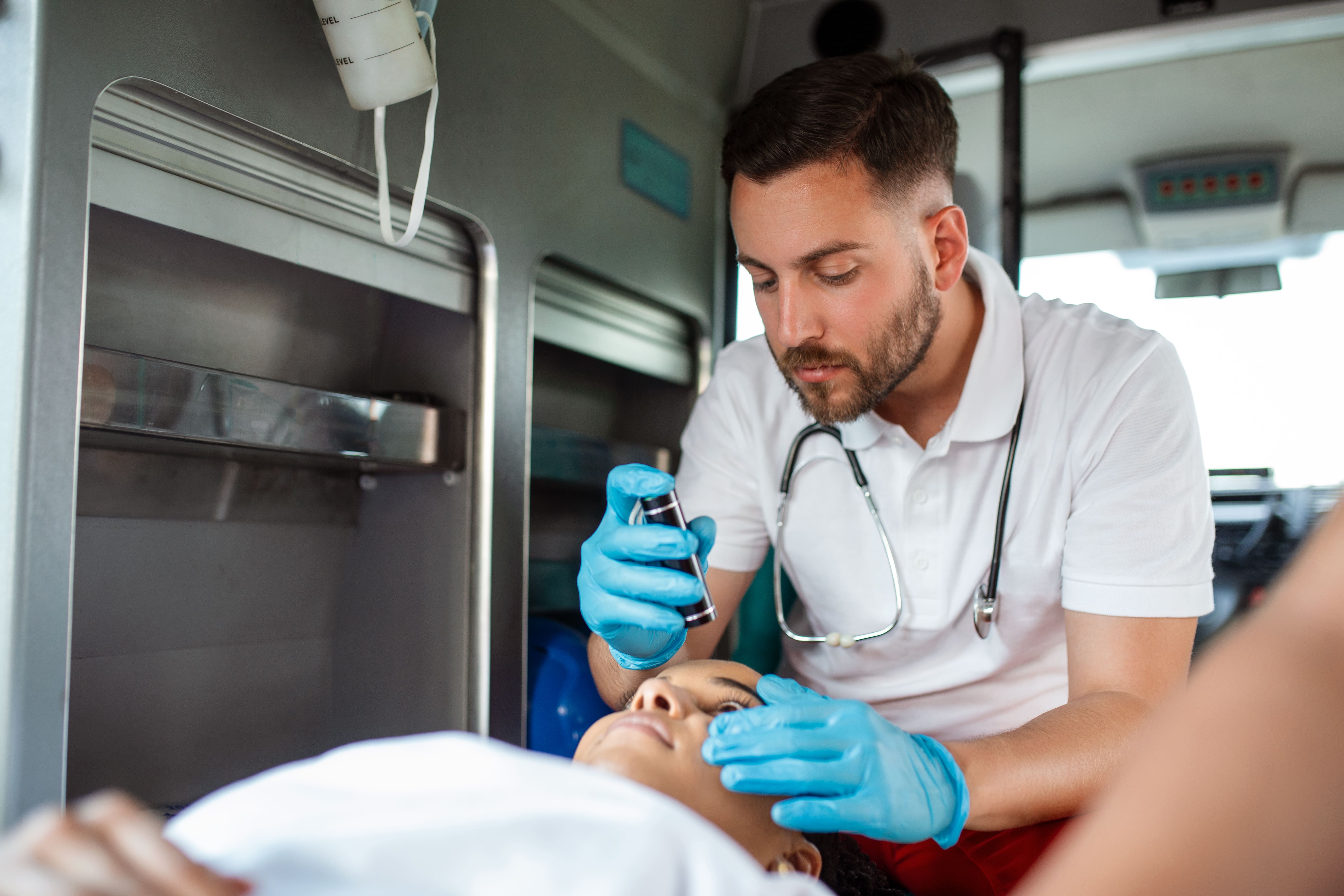 Socorrista com luvas e estetoscópio examina a pupila de uma paciente com uma lanterna dentro de uma ambulância, em atendimento de urgência.