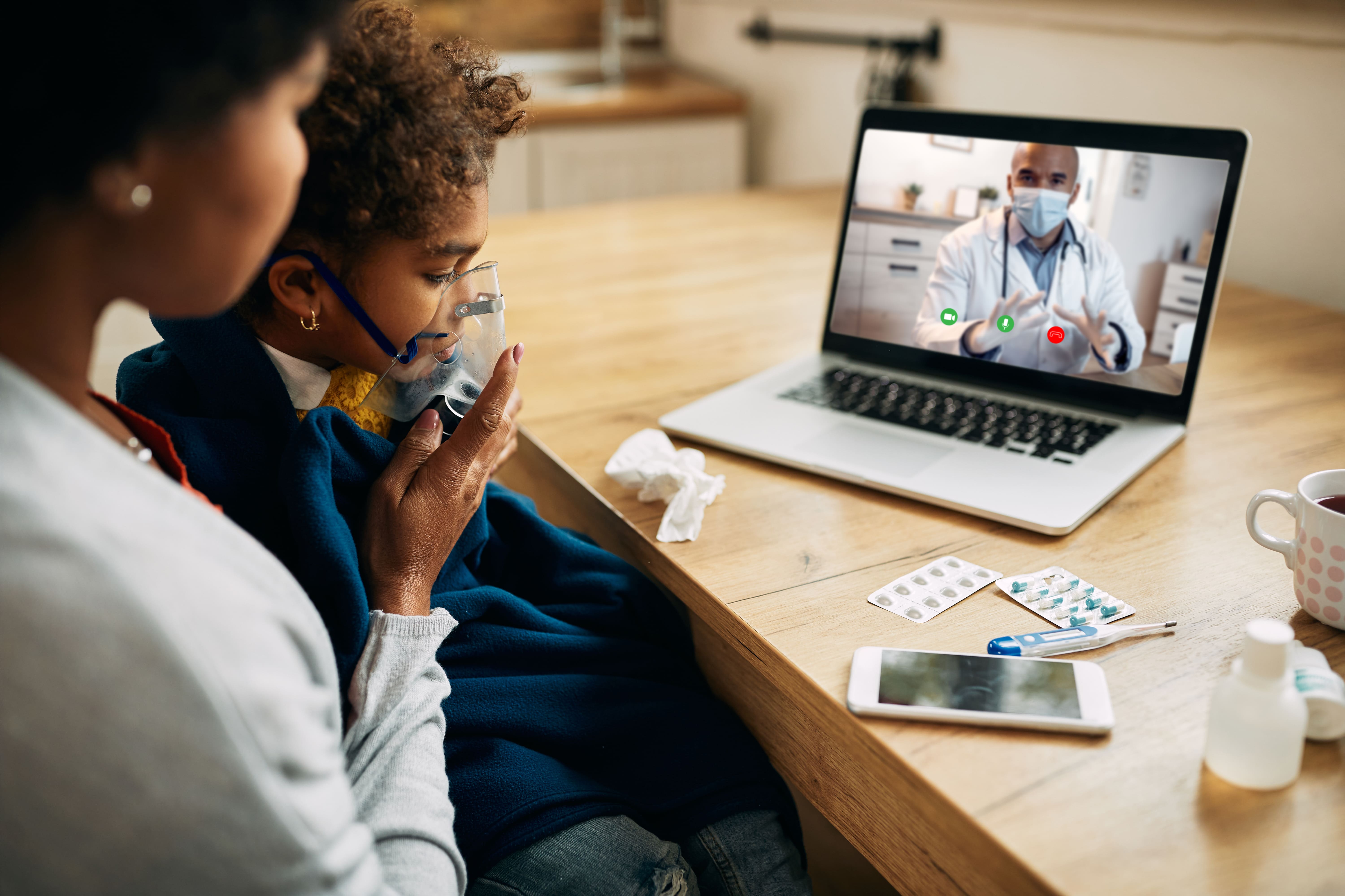 Mãe e filho fazem uma teleconsulta em casa: a criança usa máscara de inalação enquanto um médico aparece na tela do notebook.