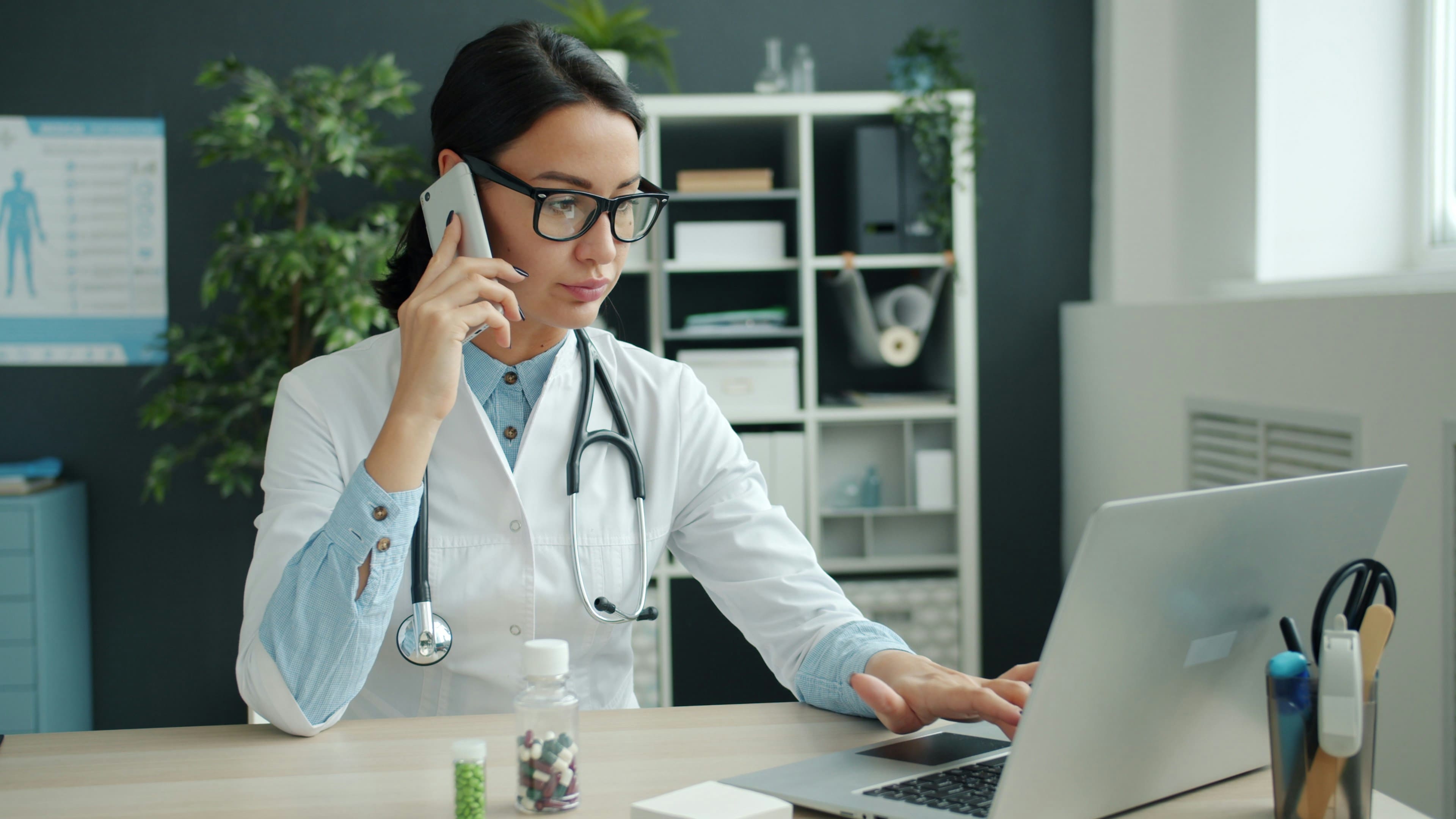 Médica de jaleco e estetoscópio fala ao celular e digita no notebook em um consultório, durante um atendimento de telemedicina.