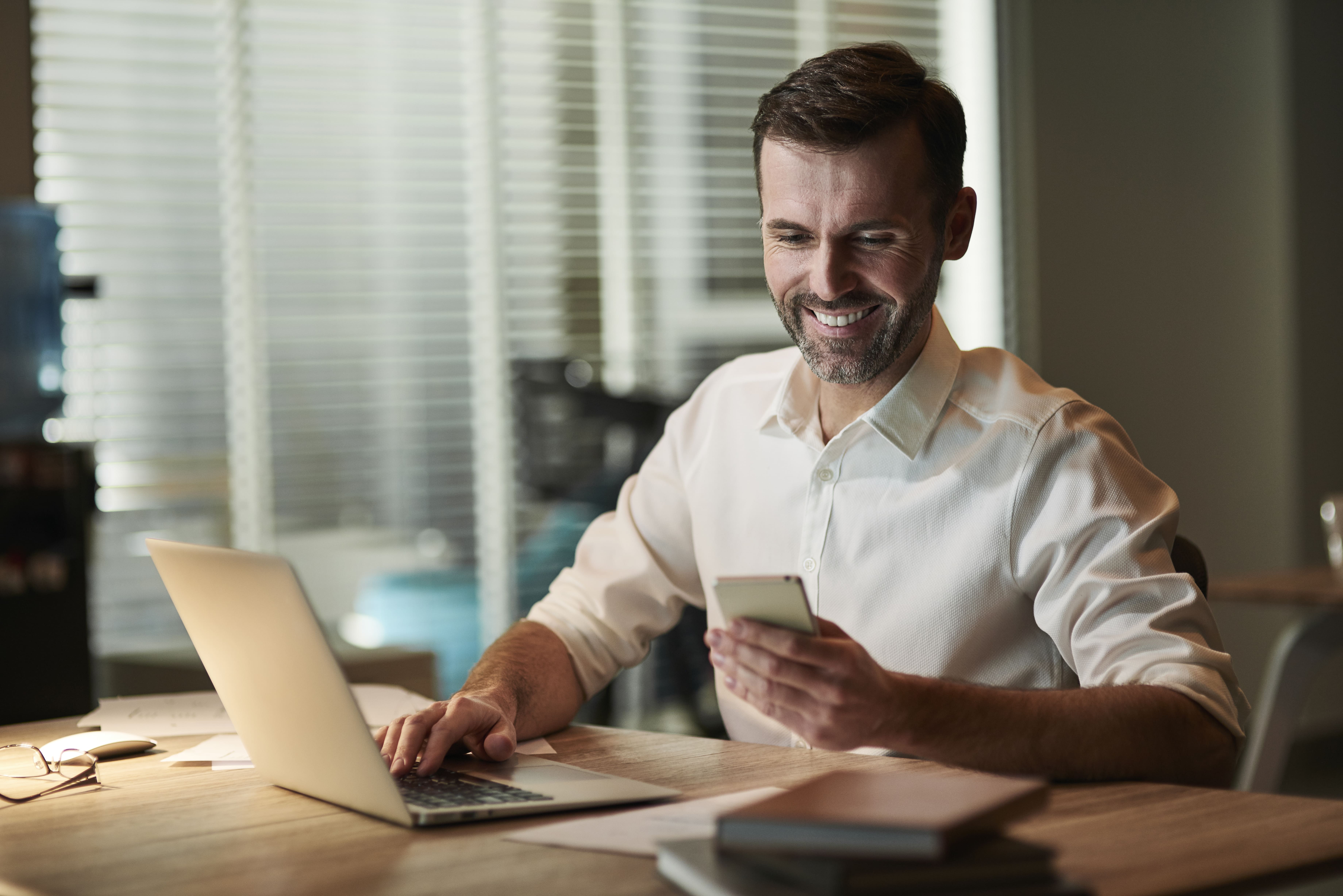 Homem sorrindo ao usar o celular e o notebook para fazer a portabilidade de carências do plano de saúde