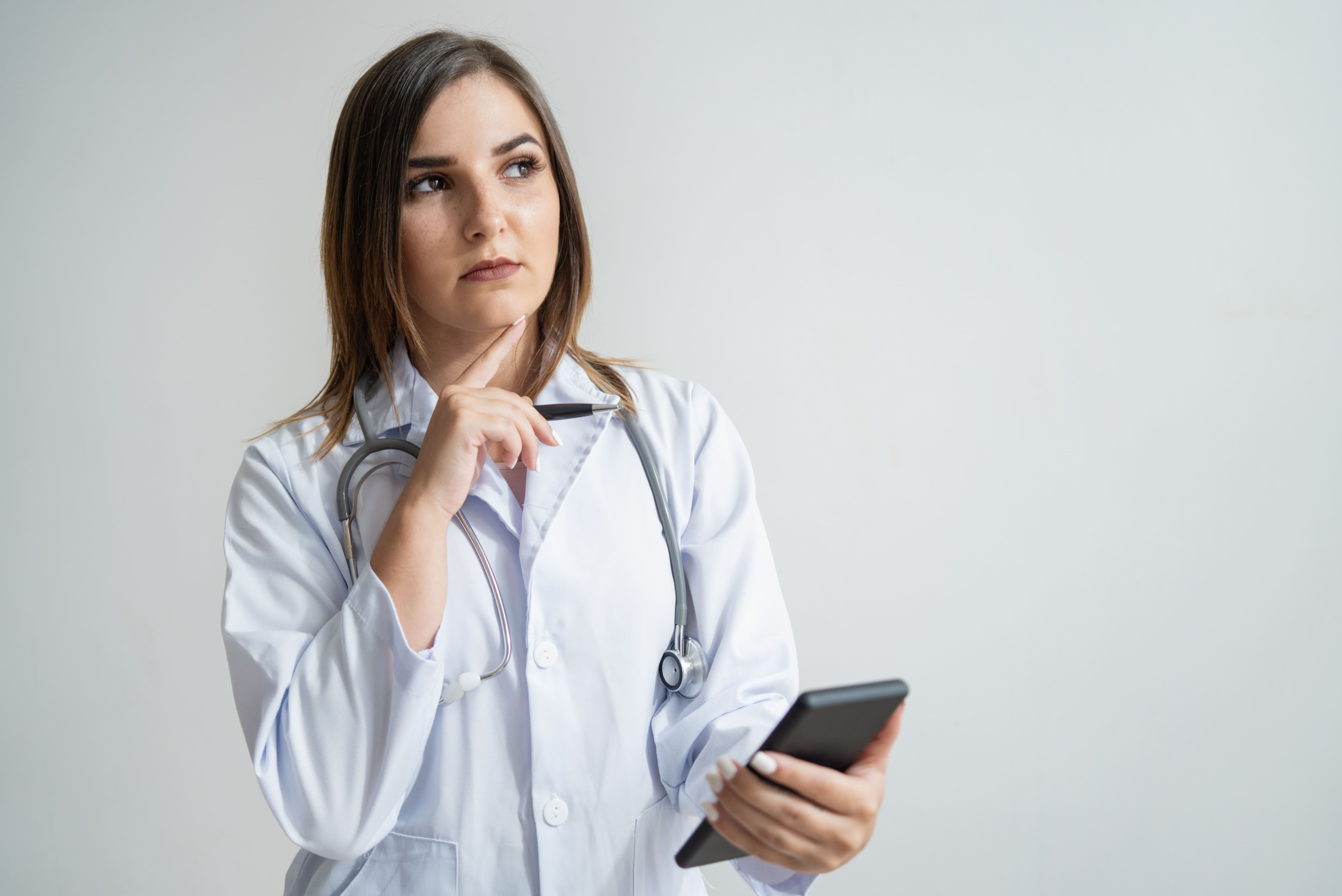 Médica de jaleco branco e estetoscópio, pensativa e com o celular na mão, avaliando qual plano de saúde escolher