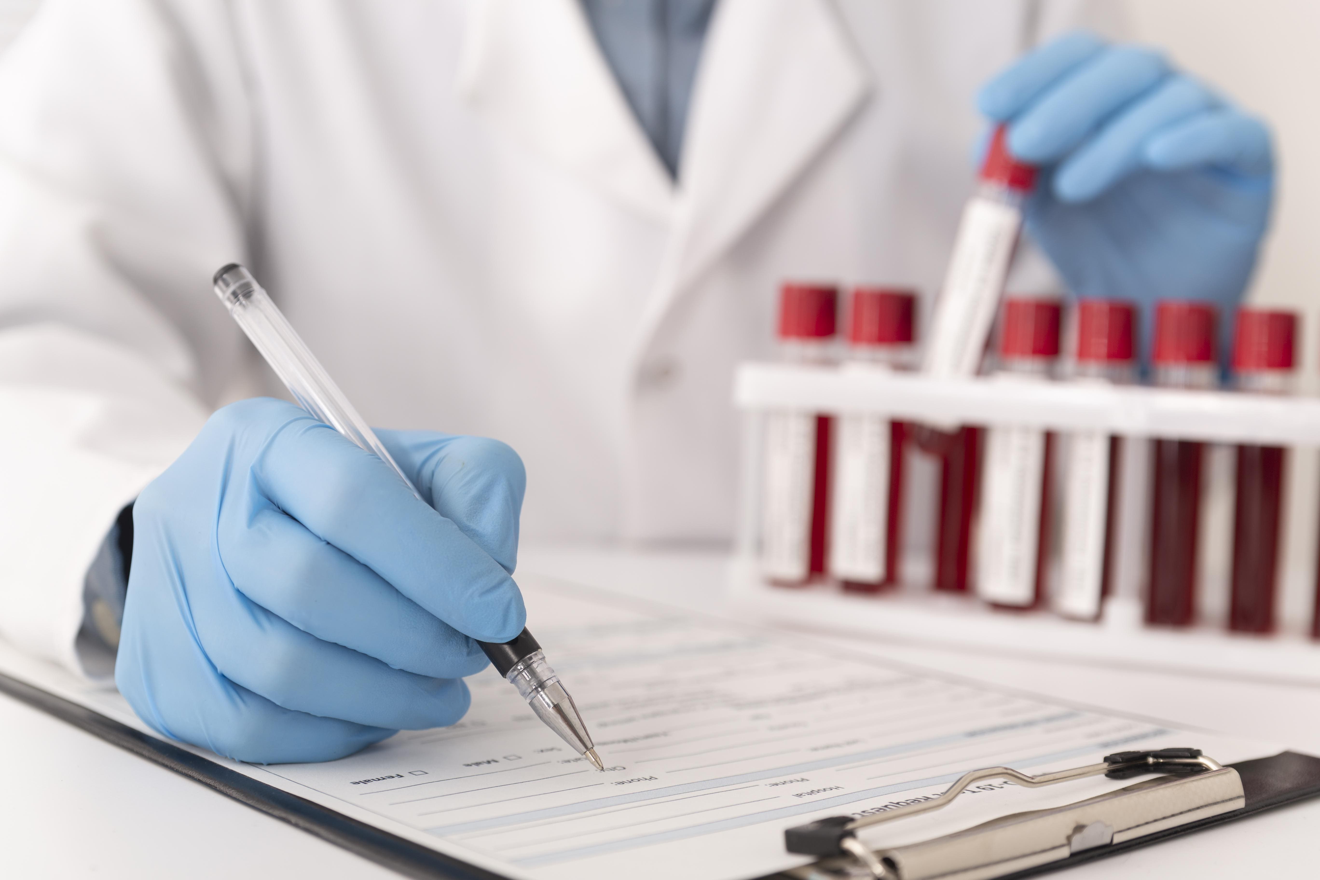 Profissional de laboratório de luvas preenchendo a requisição de um exame de sangue ao lado das amostras coletadas