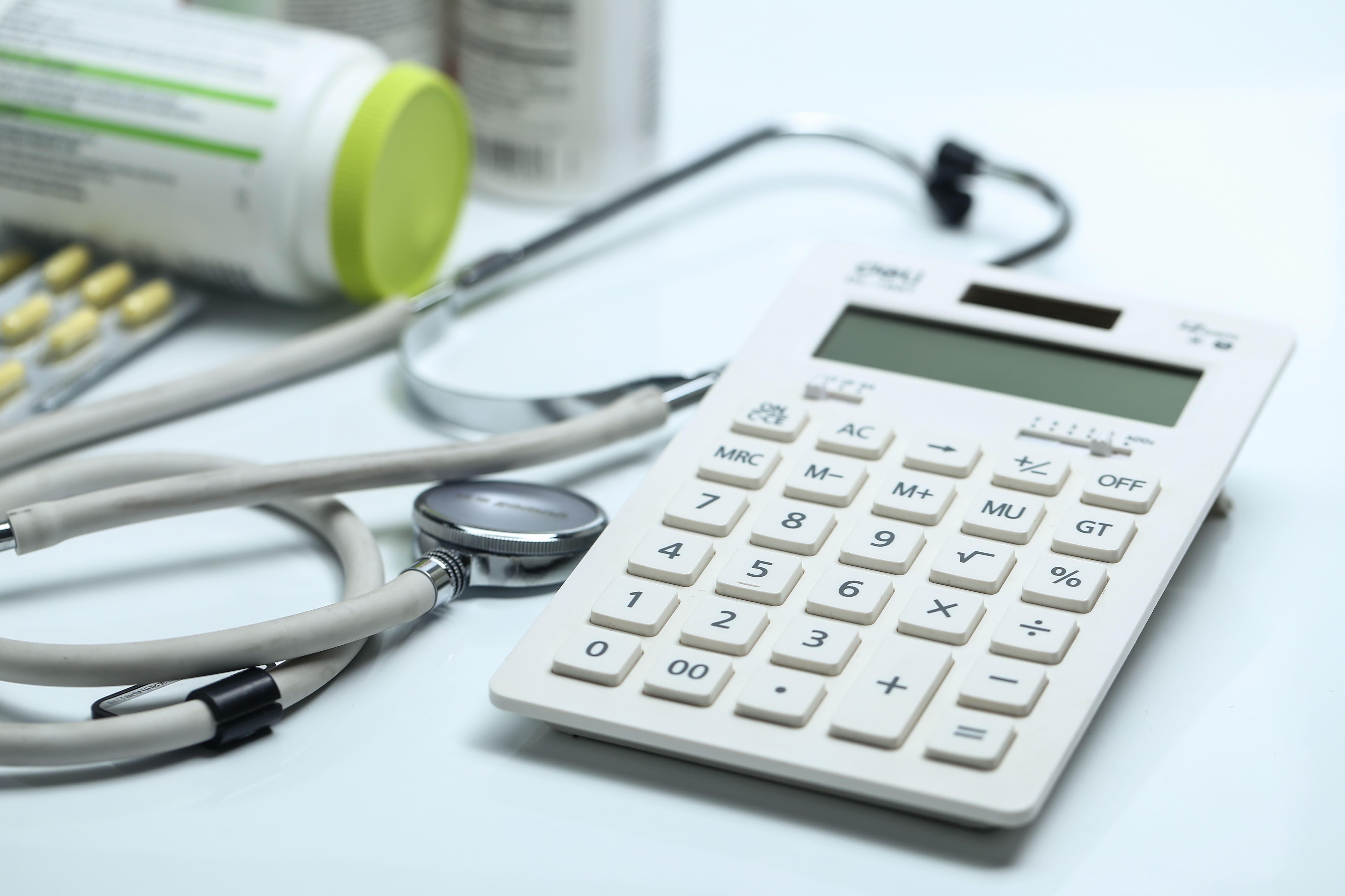 Calculadora branca ao lado de um estetoscópio e frascos de medicamentos, representando o cálculo do custo de um plano de saúde.
