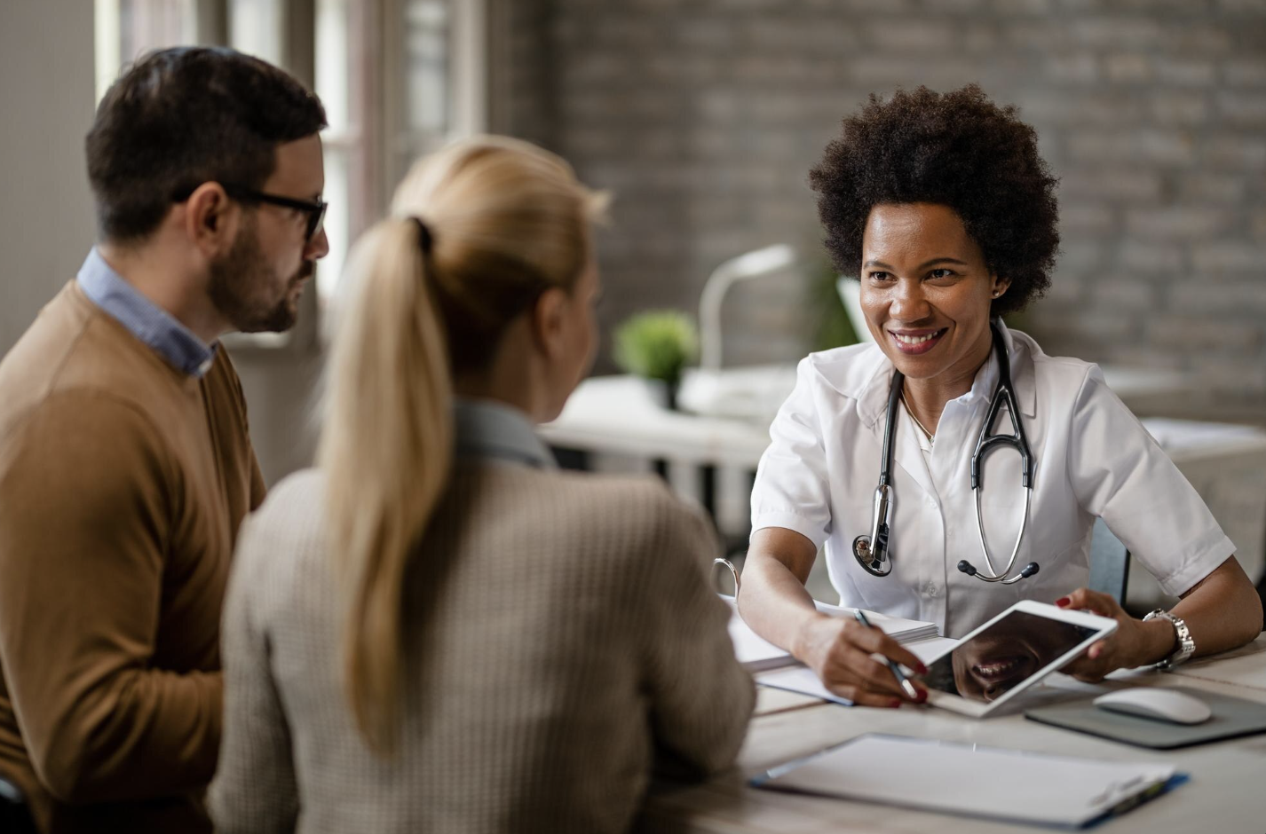 Médica de jaleco e estetoscópio conversa com um casal à mesa, mostrando informações em um tablet durante uma orientação sobre plano de saúde.