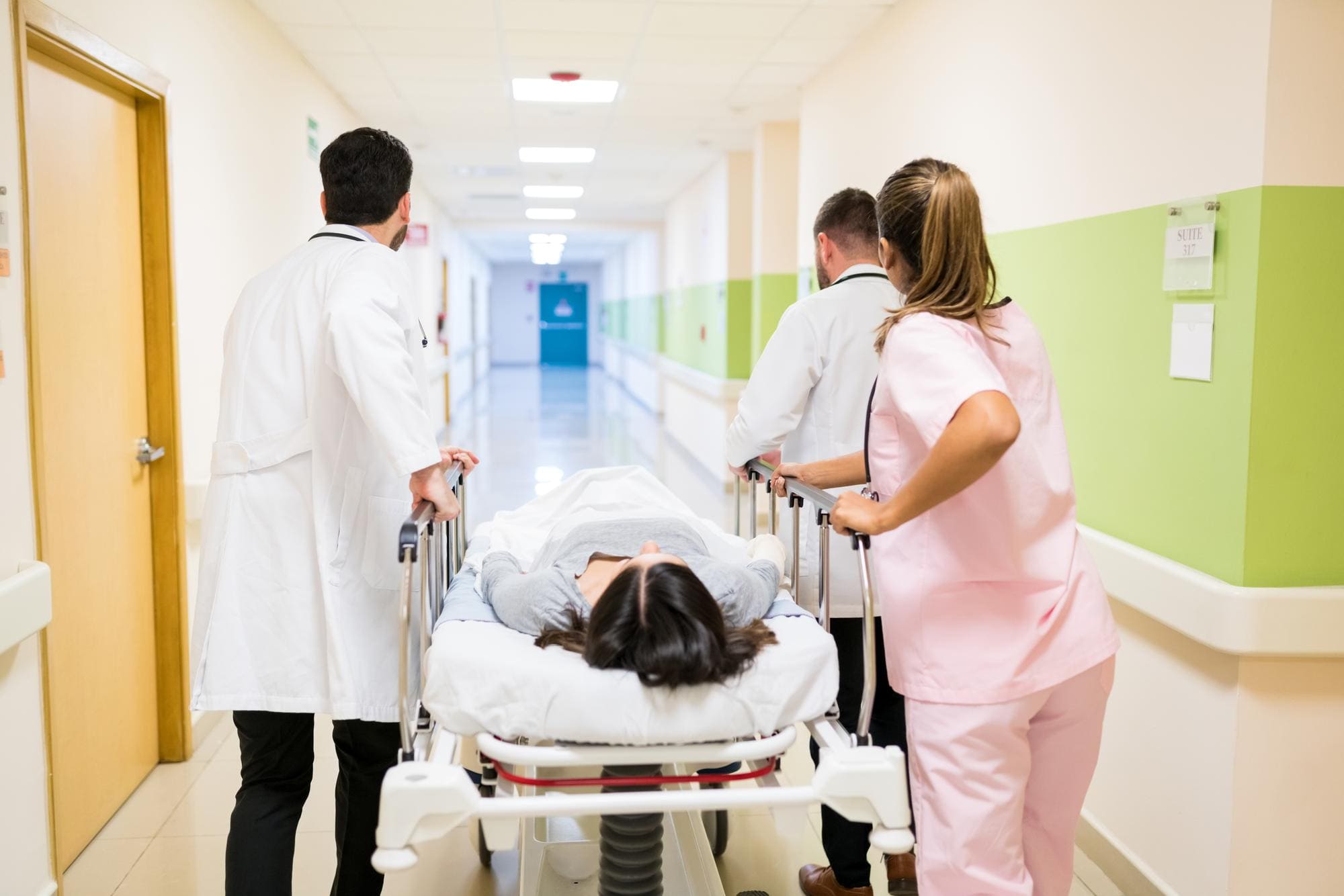 Dois médicos e uma enfermeira levam uma paciente deitada em uma maca por um corredor de hospital durante um atendimento de urgência.