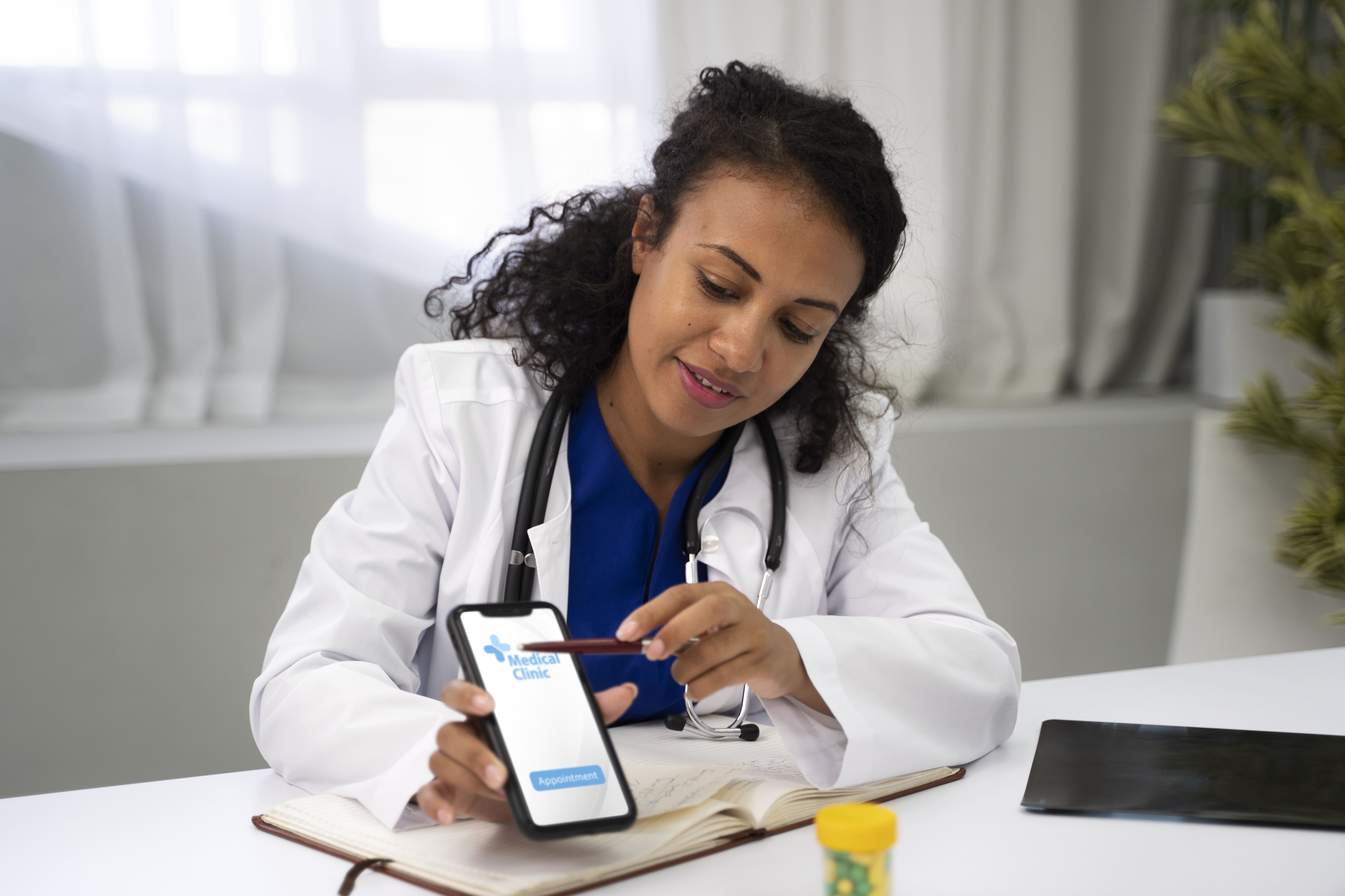 Médica de jaleco branco e estetoscópio, sentada à mesa de um consultório, mostra a tela de um aplicativo de clínica médica no celular enquanto anota em uma agenda.