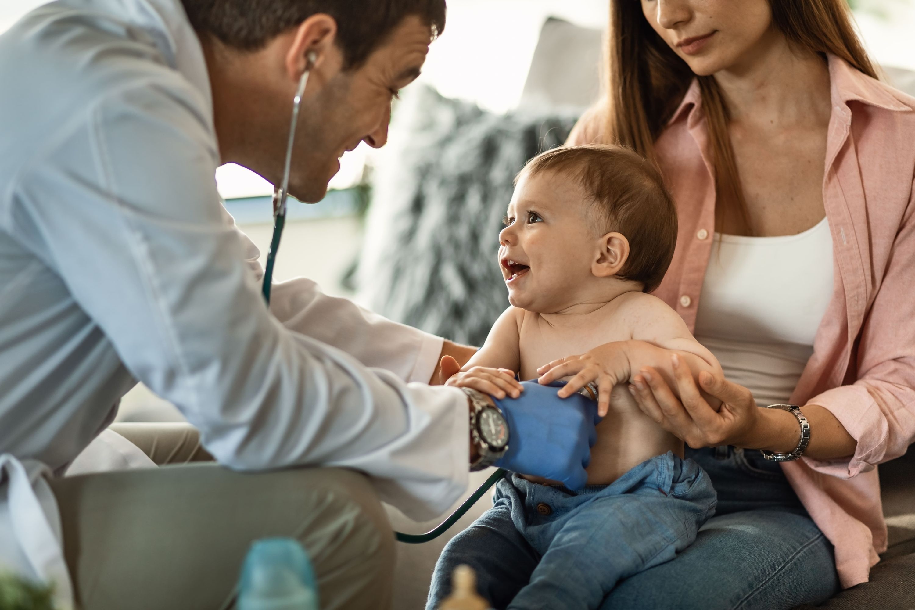 Pediatra sorridente examina um bebê sentado no colo da mãe durante uma consulta pediátrica.