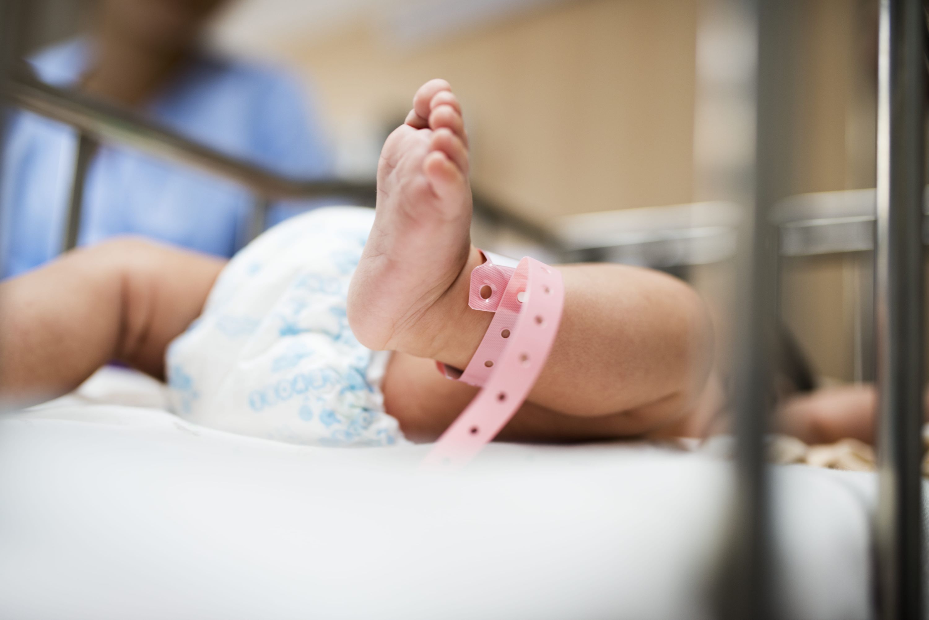 Pé de recém-nascido com pulseira de identificação em berço de maternidade, com profissional de saúde ao fundo.