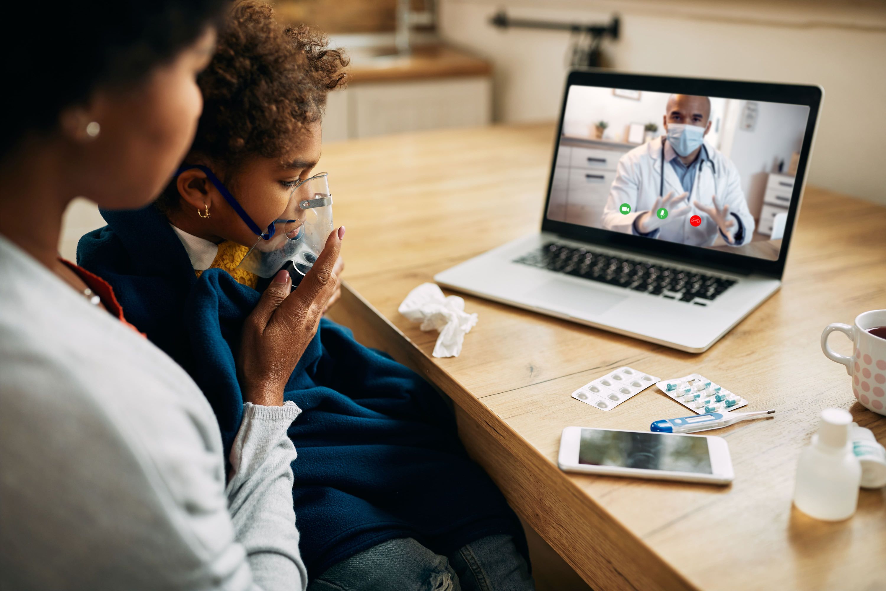 Mãe e filho fazem uma teleconsulta em casa: a criança usa máscara de inalação enquanto um médico aparece na tela do notebook.
