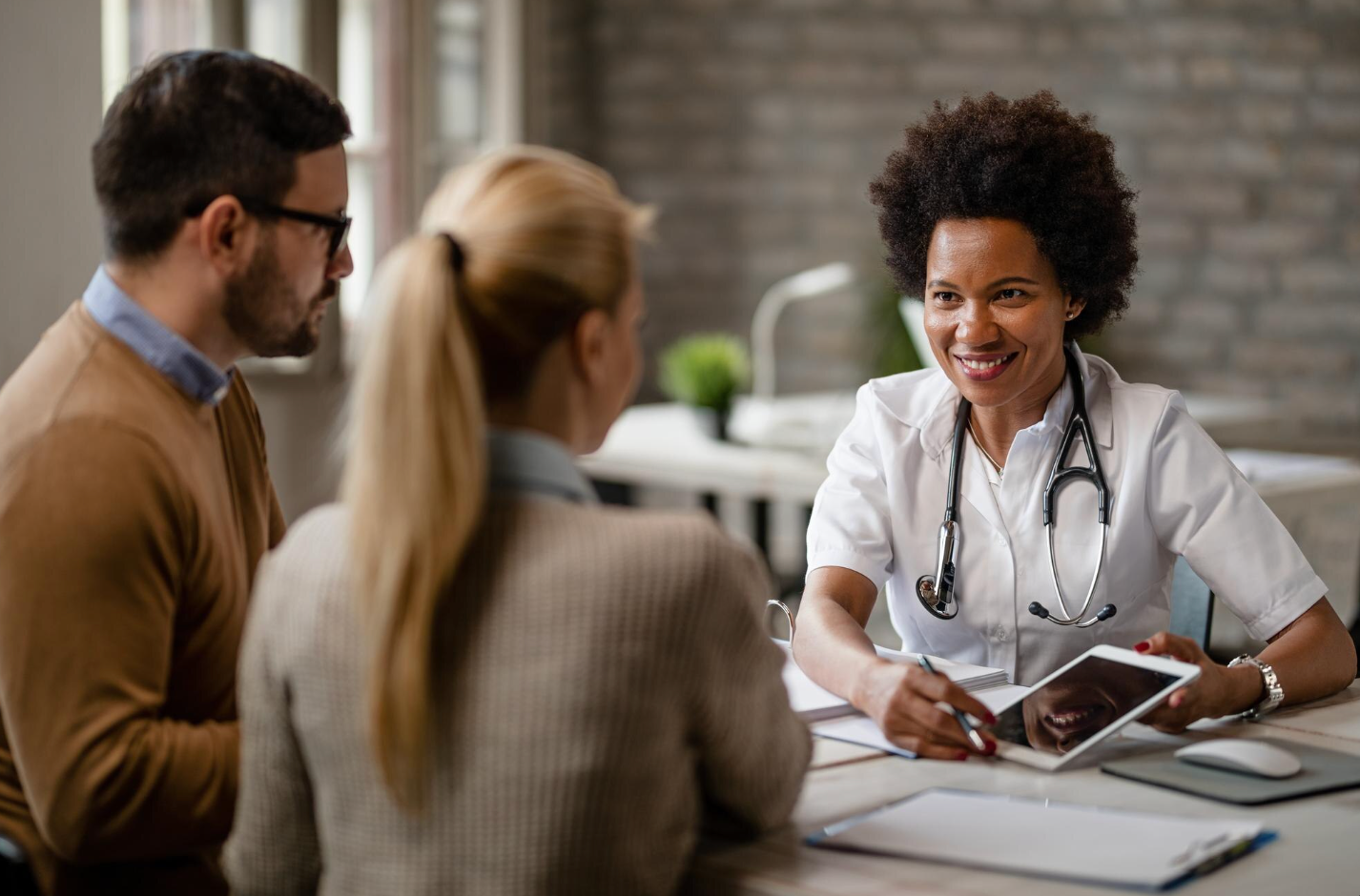 Médica de jaleco e estetoscópio conversa com um casal à mesa, mostrando informações em um tablet durante uma orientação sobre plano de saúde.